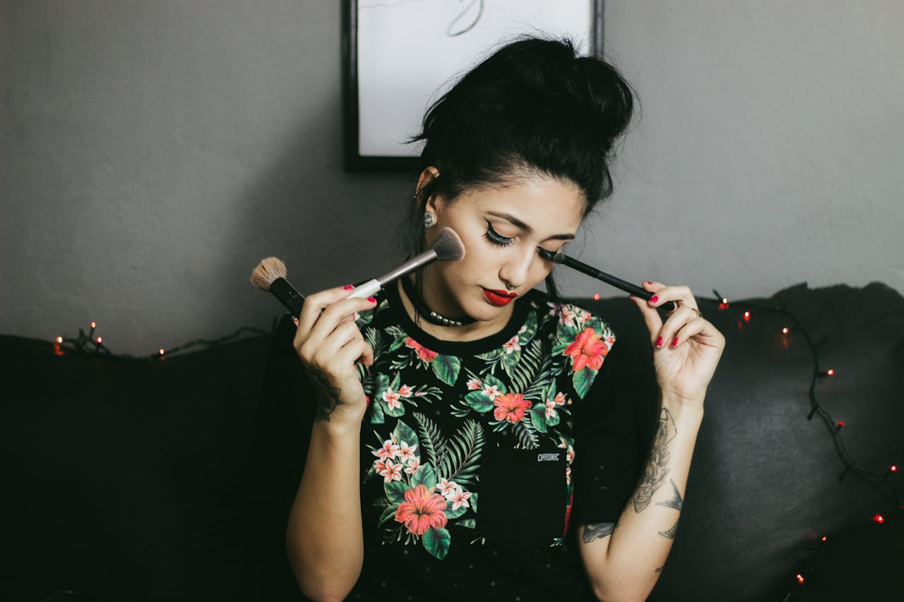 Young woman with vibrant floral shirt applying makeup using brushes indoors.