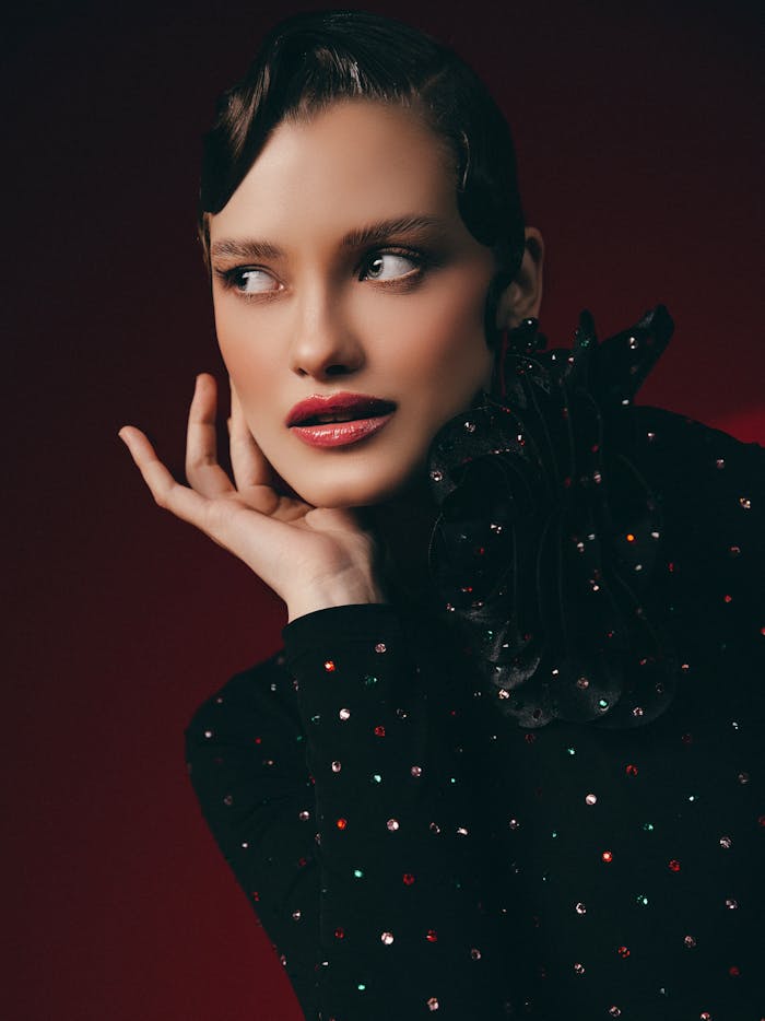 Close-up fashion portrait of a model with bold makeup and a stylish outfit against a rich red background.